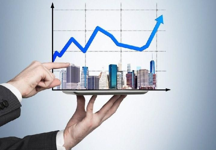 Wizualizacja wzrostu na rynku nieruchomości: ręka wskazująca na budynki na tacy, nad którymi wznosi się strzałka wykresu wzrostowego na siatce.