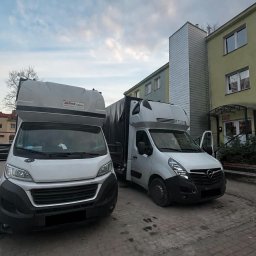 Kewin Grzybowski - Dwa busy transportowe, biały i szary, zaparkowane na brukowanym placu przed budynkiem. Jeden z busów ma otwarte drzwi.