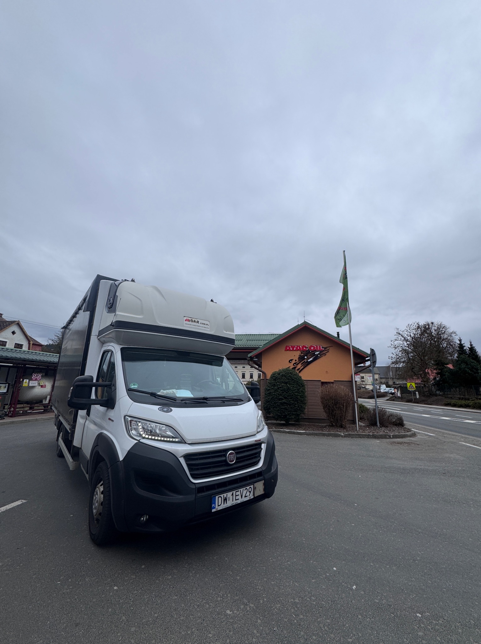 Biały bus dostawczy Fiat Ducato z wysoką zabudową, zaparkowany na parkingu przed budynkiem z logo 'ATAC OIL' w pochmurny dzień. Widoczna tablica rejestracyjna.