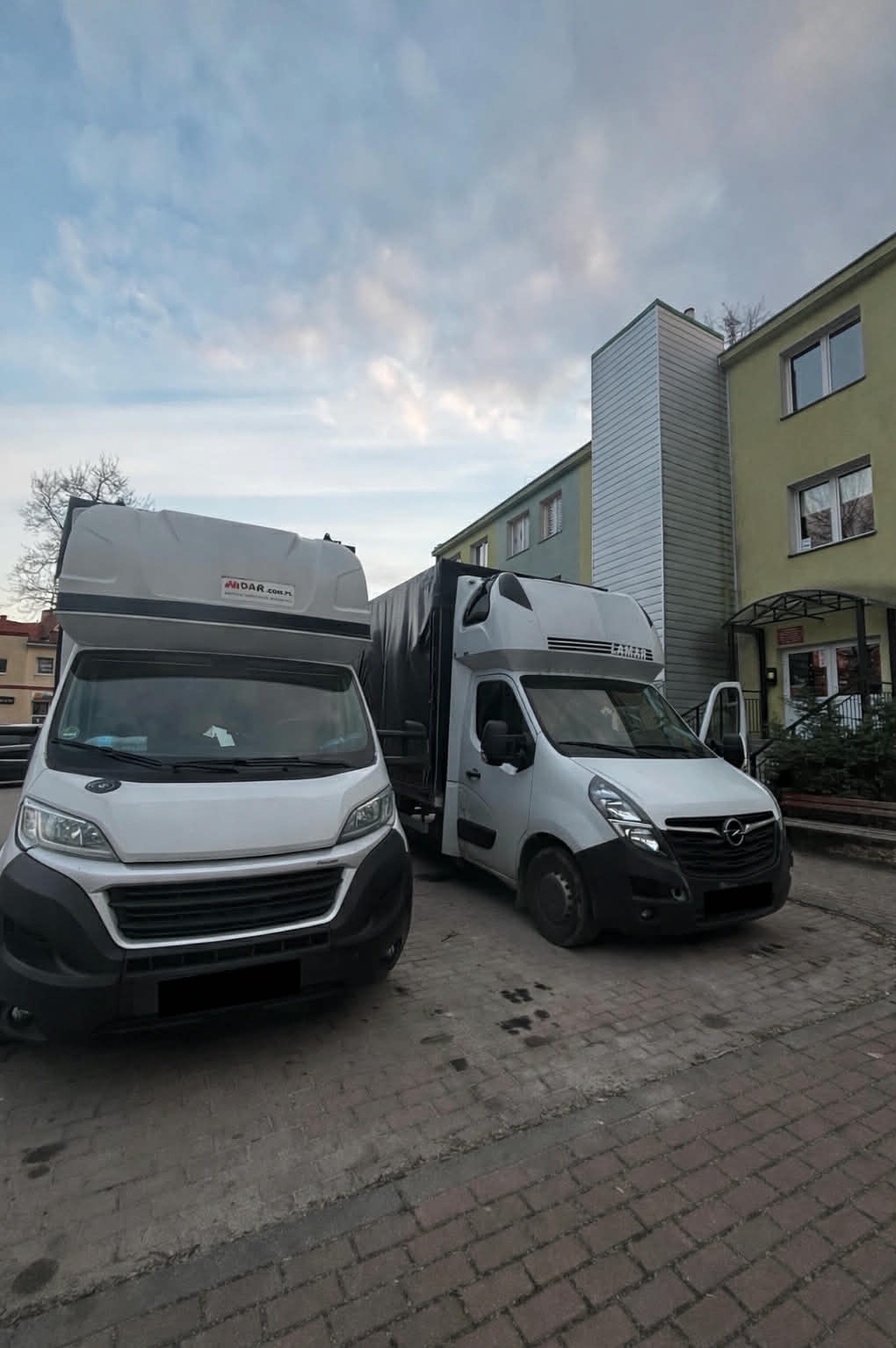 Dwa busy transportowe, biały i szary, zaparkowane na brukowanym placu przed budynkiem. Jeden z busów ma otwarte drzwi.