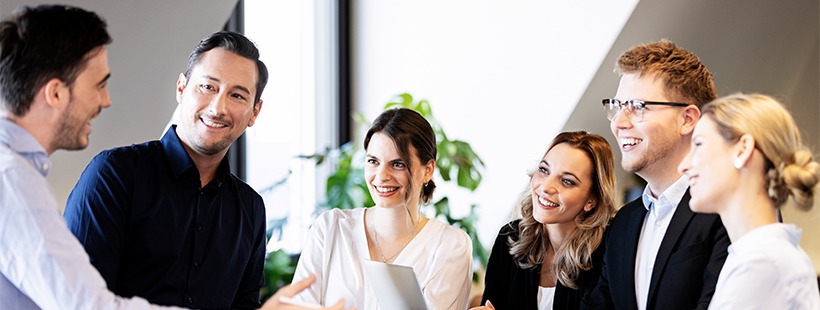 Grupa uśmiechniętych osób w biurze, w trakcie spotkania. Jasne, naturalne światło. Widoczny laptop i roślina doniczkowa w tle. Profesjonalna atmosfera.