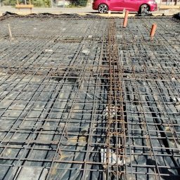 NadzórTECH Dominik Dzierwa - Zbrojenie fundamentu na czarnej folii, widoczne pionowe rury. W tle czerwony samochód. Kąt ujęcia od dołu, ukazujący siatkę zbrojeniową.