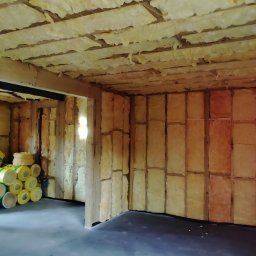 NadzórTECH Dominik Dzierwa - Wnętrze domu w trakcie izolacji termicznej: ściany i sufit wypełnione wełną mineralną, widoczne drewniane elementy konstrukcyjne oraz rolki izolacji w tle.