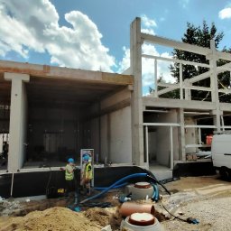 NadzórTECH Dominik Dzierwa - Budowa domu w stanie surowym otwartym, widoczni dwaj pracownicy w kaskach i kamizelkach odblaskowych, zaparkowany biały bus. Elementy kanalizacji na pierwszym planie.