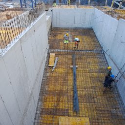 NadzórTECH Dominik Dzierwa - Zalewanie fundamentów: betonowe ściany szalunkowe, zbrojenie stalowe. Robotnicy w kaskach nadzorują prace. Widok z góry na plac budowy, etap wylewania fundamentów.