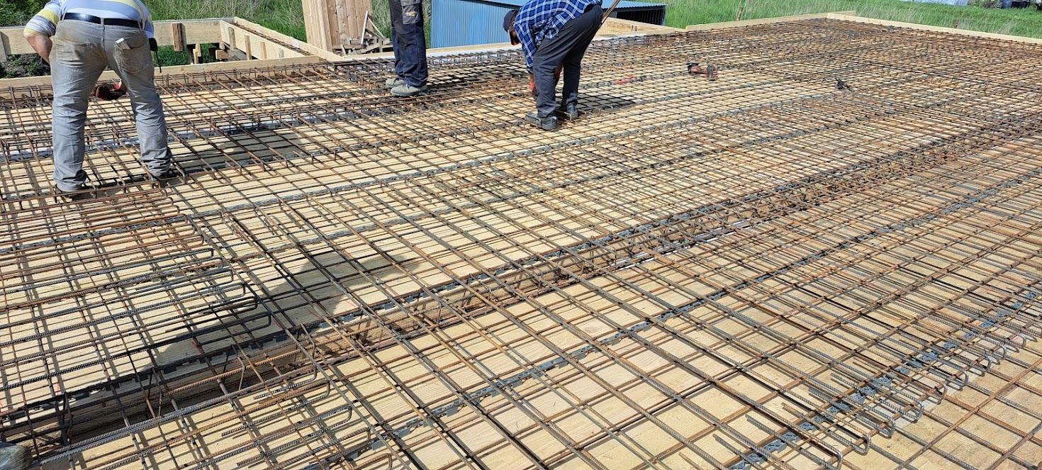 Zbrojenie stropu na deskowaniu, widoczne pręty stalowe tworzące siatkę, elementy dystansowe i fragmenty sylwetek osób wykonujących prace. Jasne oświetlenie dzienne.