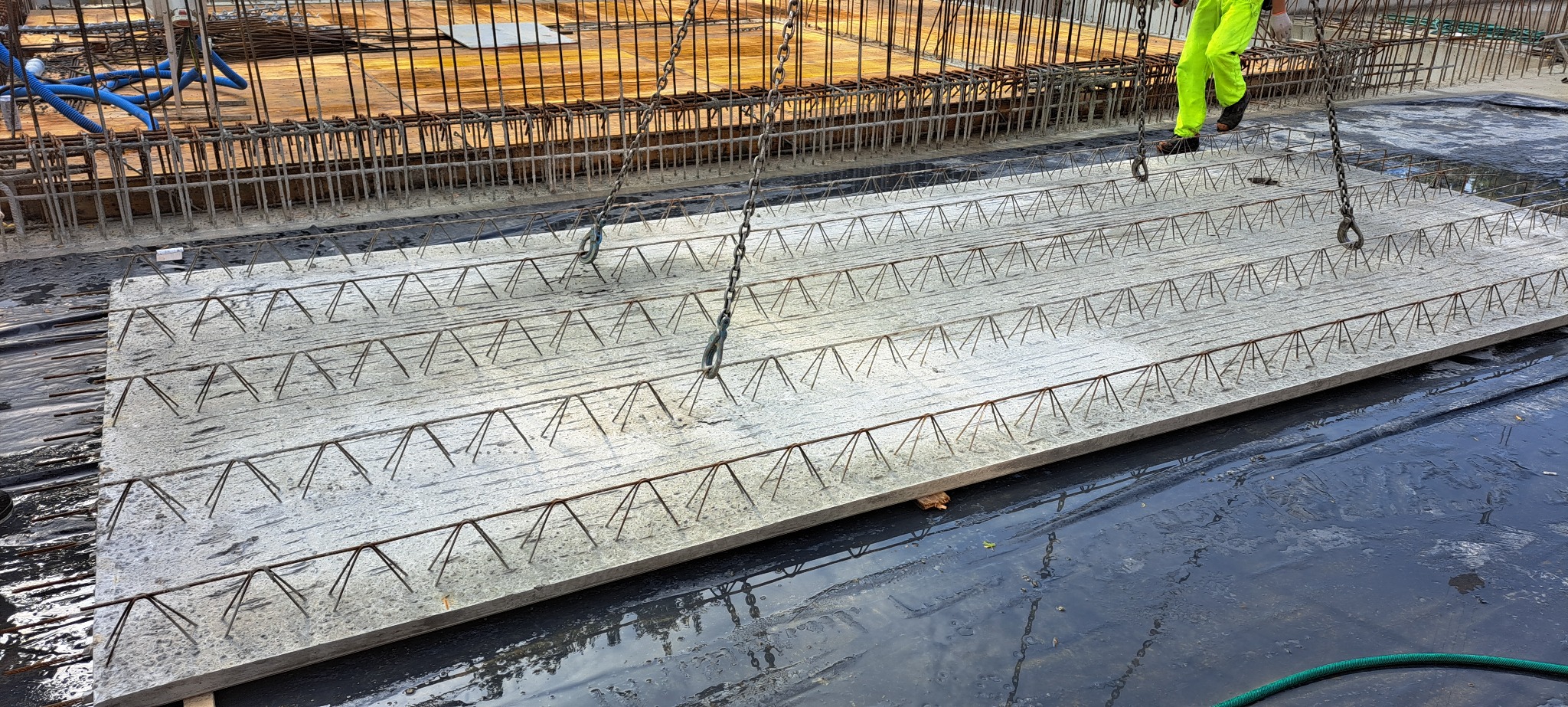 Prefabrykowany strop żelbetowy z wystającymi prętami, podnoszony na budowie za pomocą łańcuchów, w tle widoczne zbrojenie i szalunki.
