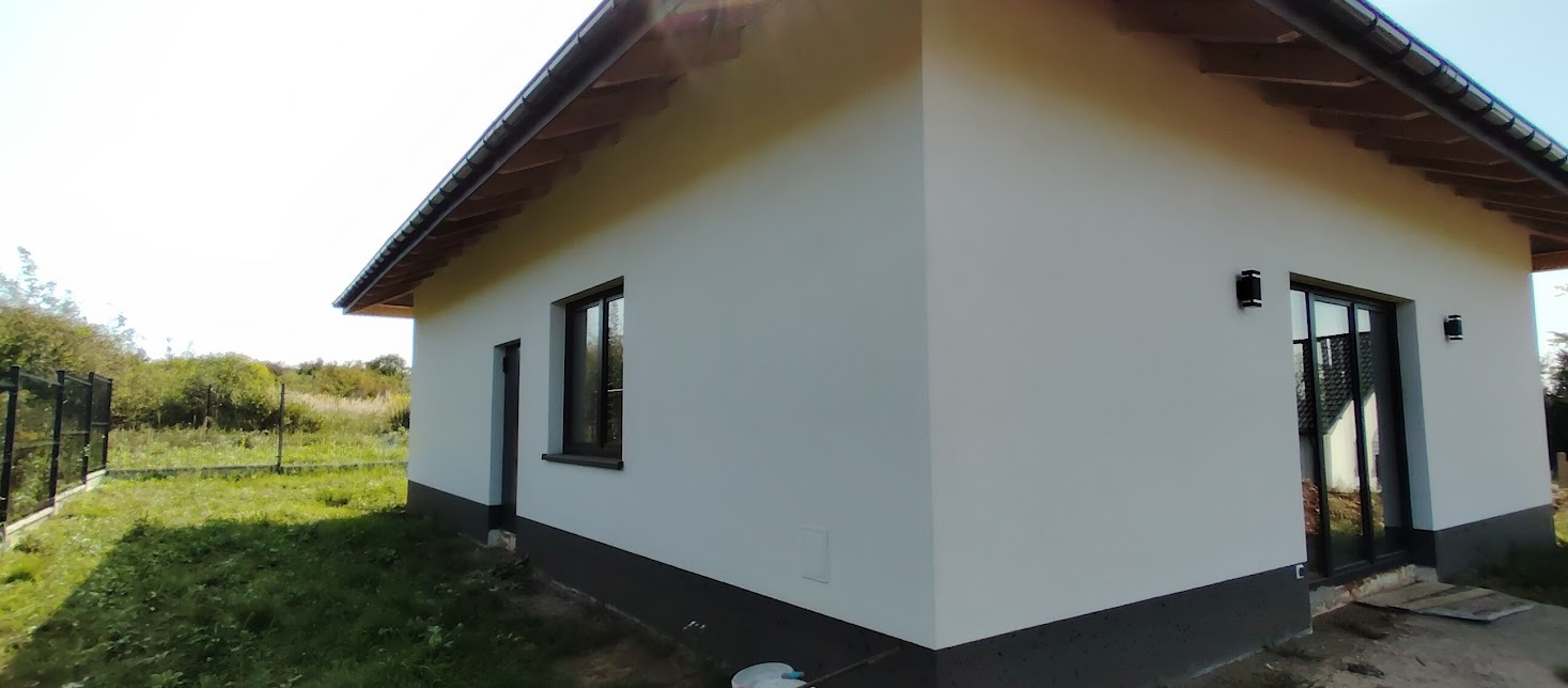 Biały parterowy dom z ciemną podmurówką i drewnianym dachem, widziany z zewnątrz, na tle zieleni. Widoczne okna, drzwi przesuwne i oświetlenie zewnętrzne.