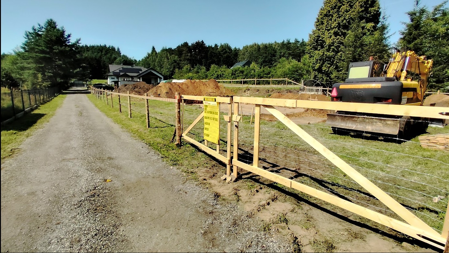 Teren budowy z wykopem, ogrodzony drewnianą bramą i siatką. W tle dom z ciemnym dachem i żółta koparka. Tablica informacyjna na bramie. Perspektywa z drogi dojazdowej.