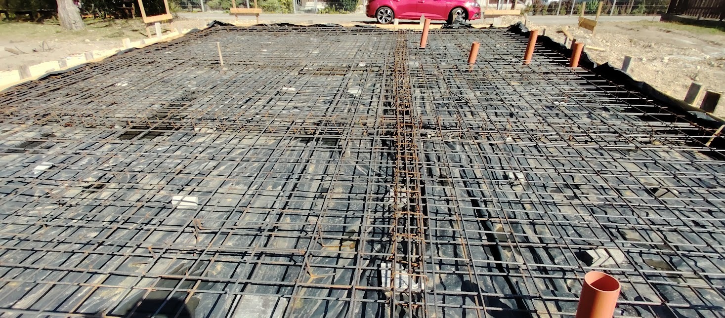 Zbrojenie fundamentu na czarnej folii, widoczne pionowe rury. W tle czerwony samochód. Kąt ujęcia od dołu, ukazujący siatkę zbrojeniową.