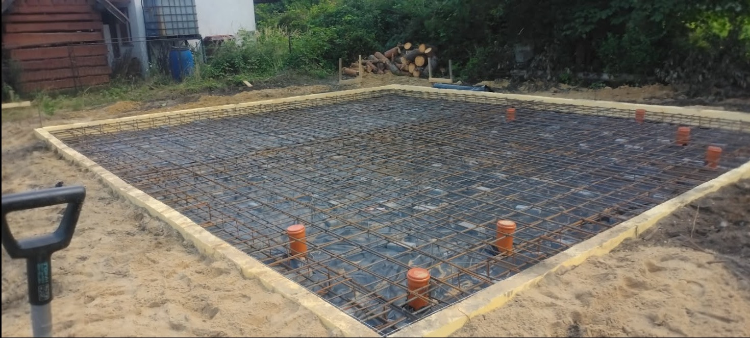 Zbrojenie fundamentu pod budowę domu jednorodzinnego z widocznymi rurami kanalizacyjnymi i deskowaniem. W tle drzewa i zbiornik na wodę.