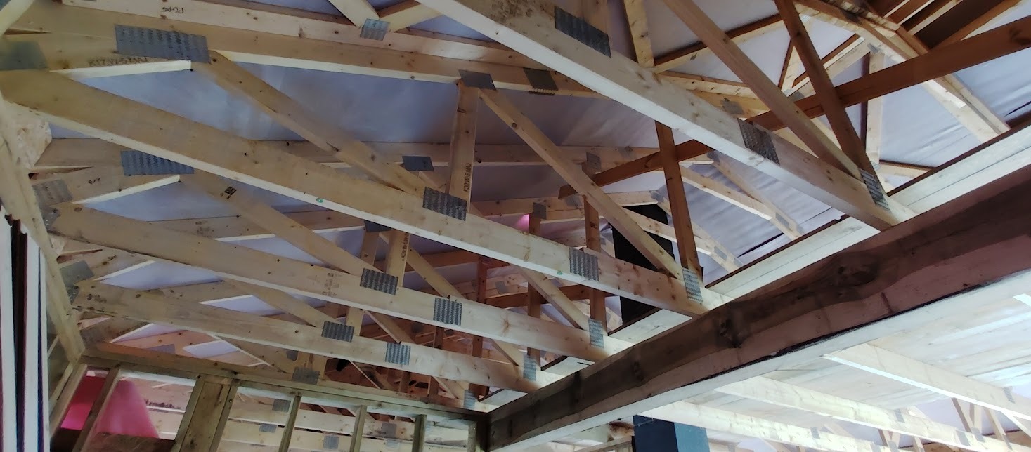 Konstrukcja drewniana dachu w trakcie budowy, widoczne elementy więźby z metalowymi łącznikami. Jasne drewno, biała membrana dachowa. Ujęcie z dołu.