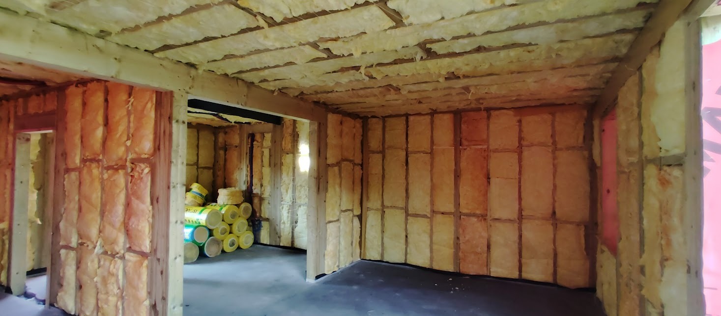 Wnętrze domu w trakcie izolacji termicznej: ściany i sufit wypełnione wełną mineralną, widoczne drewniane elementy konstrukcyjne oraz rolki izolacji w tle.