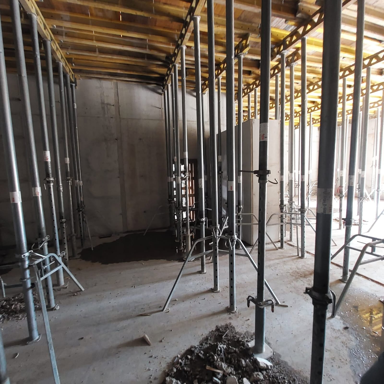 Wnętrze w trakcie budowy z podporami stropowymi i szalunkiem. Widoczne metalowe stemple i drewniane elementy konstrukcyjne, surowy beton na ścianach i podłodze.