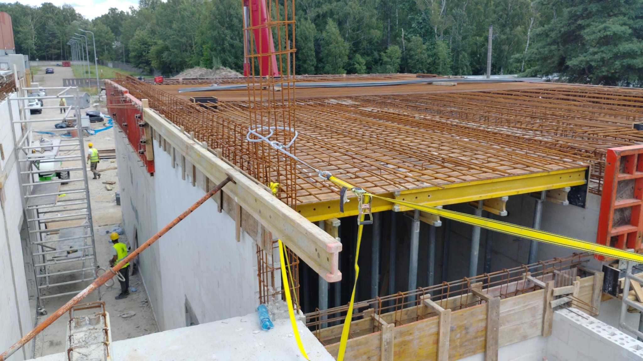 Zbrojenie stropu budynku w trakcie budowy, widoczne stalowe pręty, szalunki i pracownicy w kaskach. Ujęcie z góry, z widocznym lasem w tle.