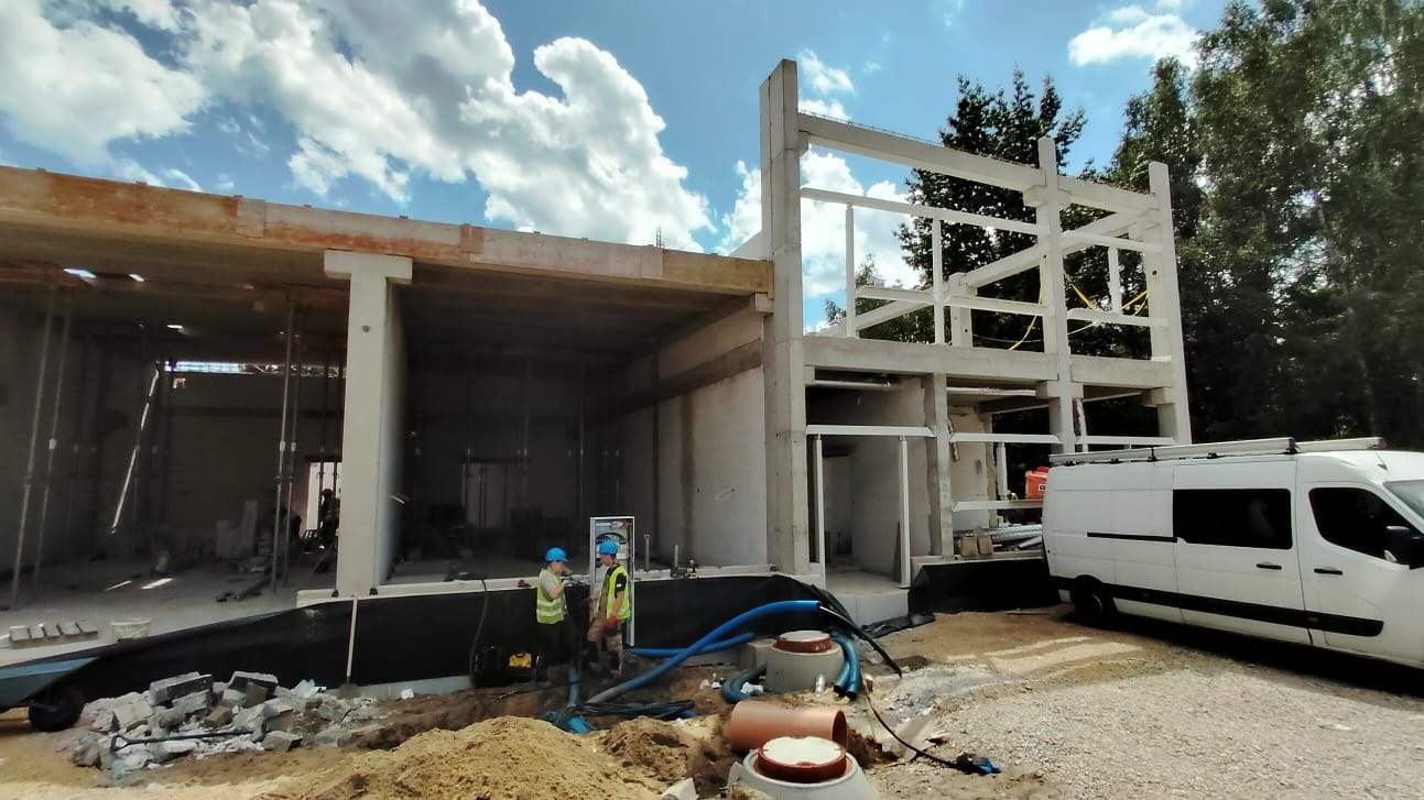 Budowa domu w stanie surowym otwartym, widoczni dwaj pracownicy w kaskach i kamizelkach odblaskowych, zaparkowany biały bus. Elementy kanalizacji na pierwszym planie.