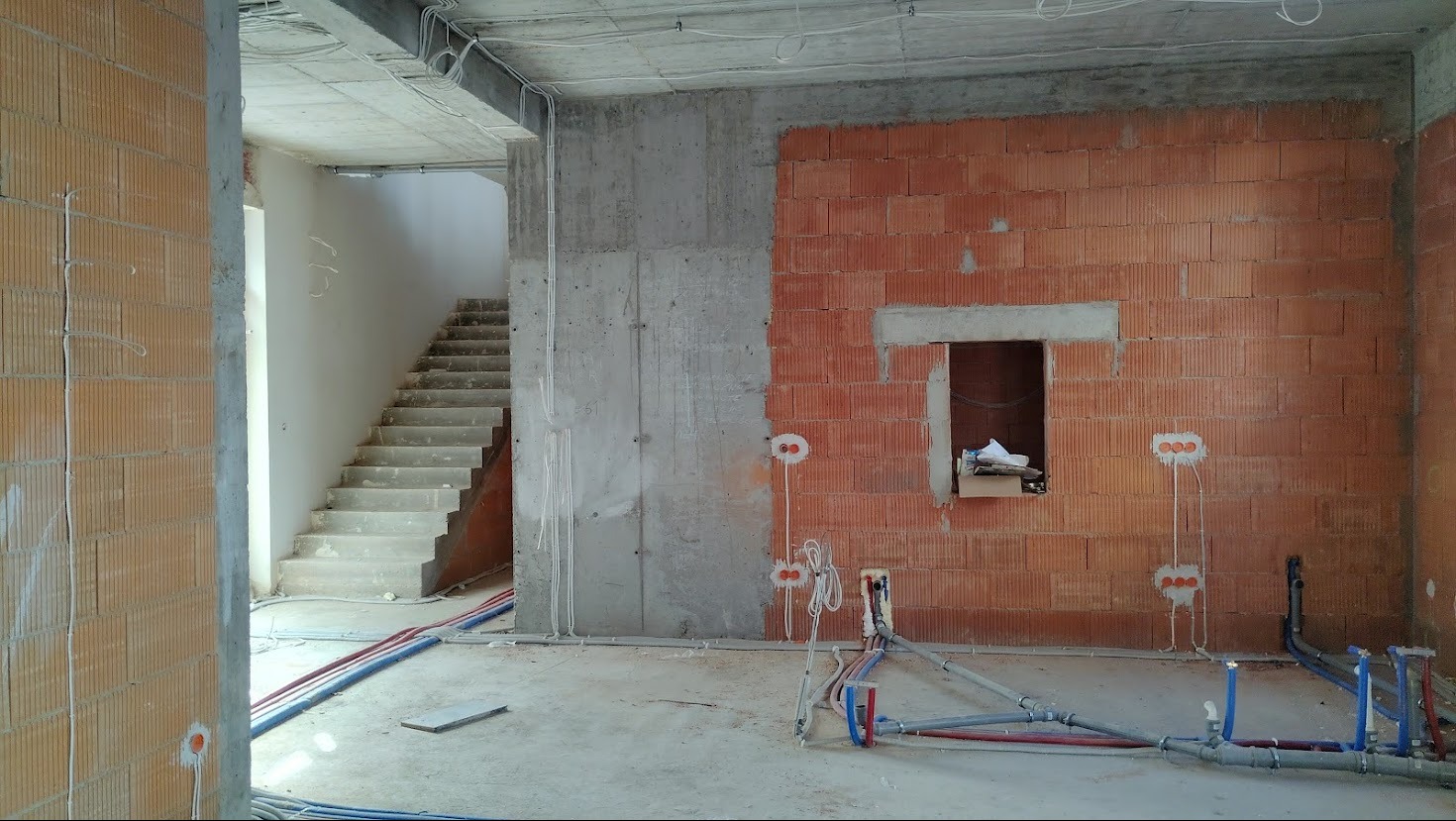 Surowy stan wnętrza: ściana z cegły, beton, instalacje elektryczne i hydrauliczne, schody w tle. Wnętrze w trakcie prac wykończeniowych.