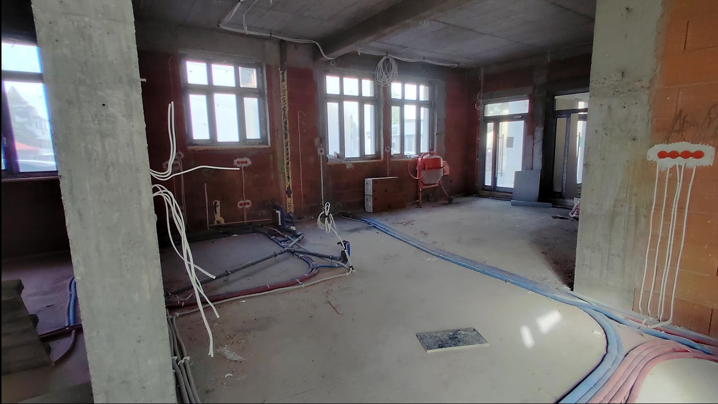 Wnętrze w stanie surowym: widoczne instalacje elektryczne i hydrauliczne na ścianach z cegły i betonowych kolumnach. W tle okna i betonowa podłoga.
