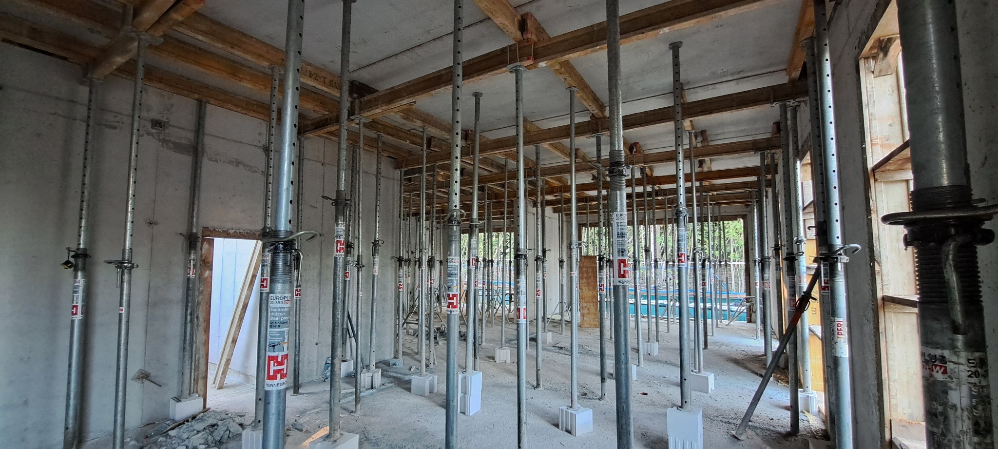 Wnętrze w trakcie budowy: betonowe ściany, drewniane belki stropowe i liczne metalowe stemple podpierające strop. Widok na zewnątrz przez otwory okienne.