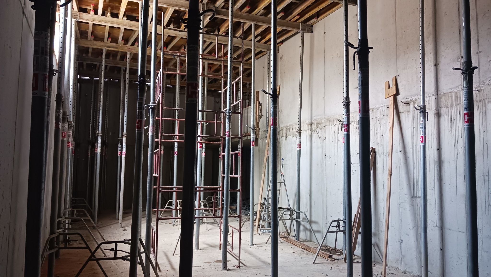 Wnętrze w trakcie budowy: betonowe ściany, stalowe podpory stropowe z regulacją wysokości, widoczne drewniane szalunki stropu. Etap wylewania stropu.