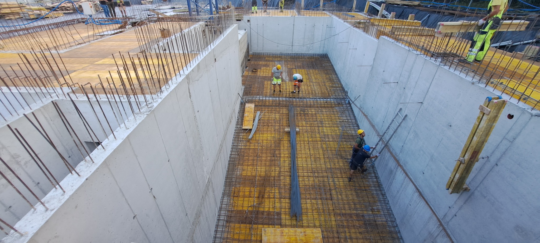 Zalewanie fundamentów: betonowe ściany szalunkowe, zbrojenie stalowe. Robotnicy w kaskach nadzorują prace. Widok z góry na plac budowy, etap wylewania fundamentów.