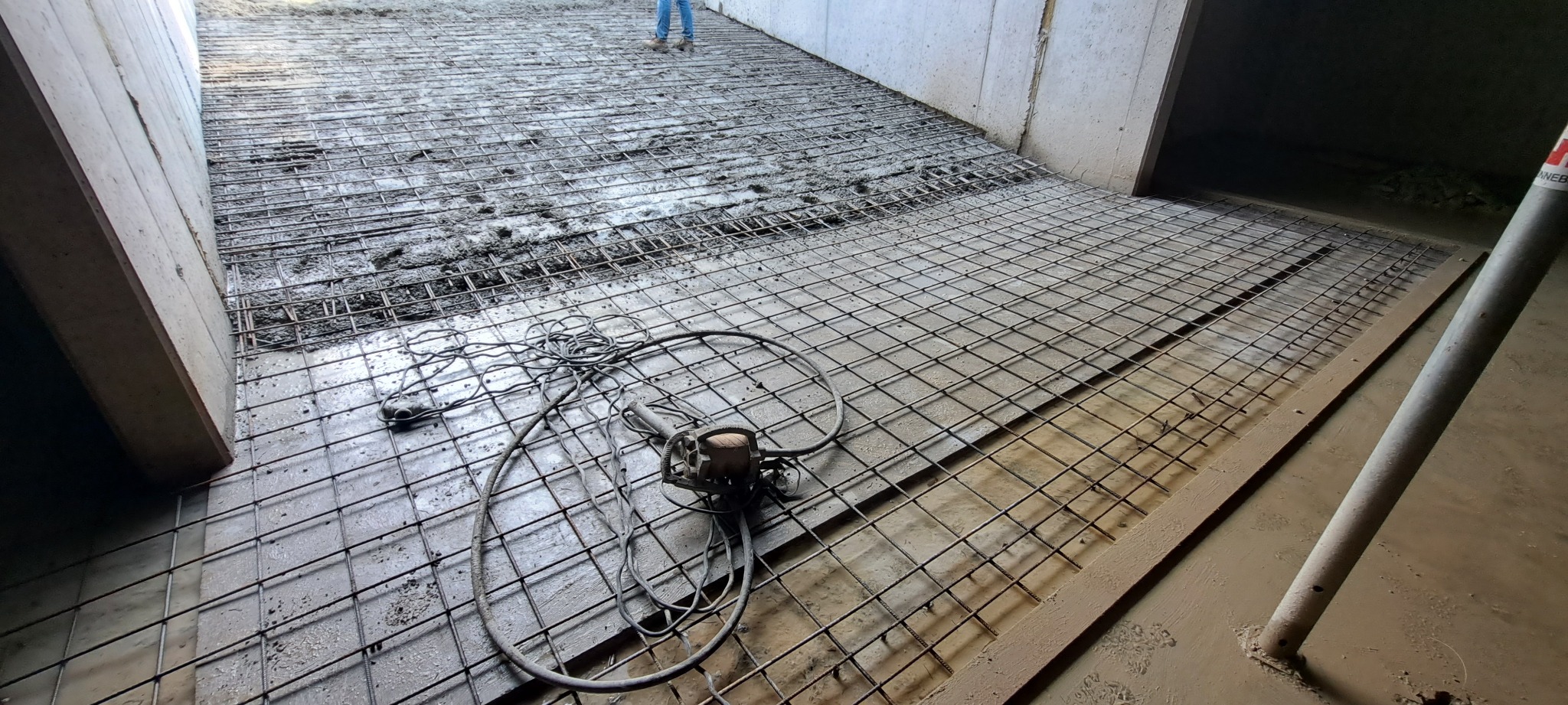 Zbrojenie pod wylewkę betonową w hali. Widoczna siatka zbrojeniowa, częściowo zalana betonem i narzędzie do wiązania drutu. Prace budowlane w surowym wnętrzu.