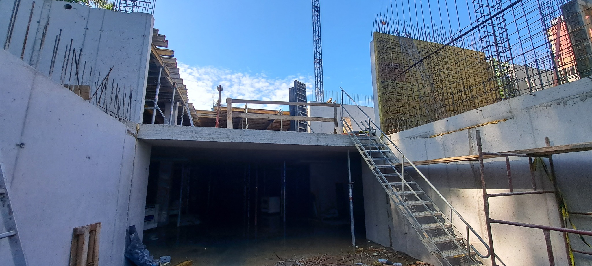 Surowy stan budowy: betonowe ściany, zbrojenia, szalunki i metalowe schody na tle błękitnego nieba. Widoczny dźwig budowlany w oddali.