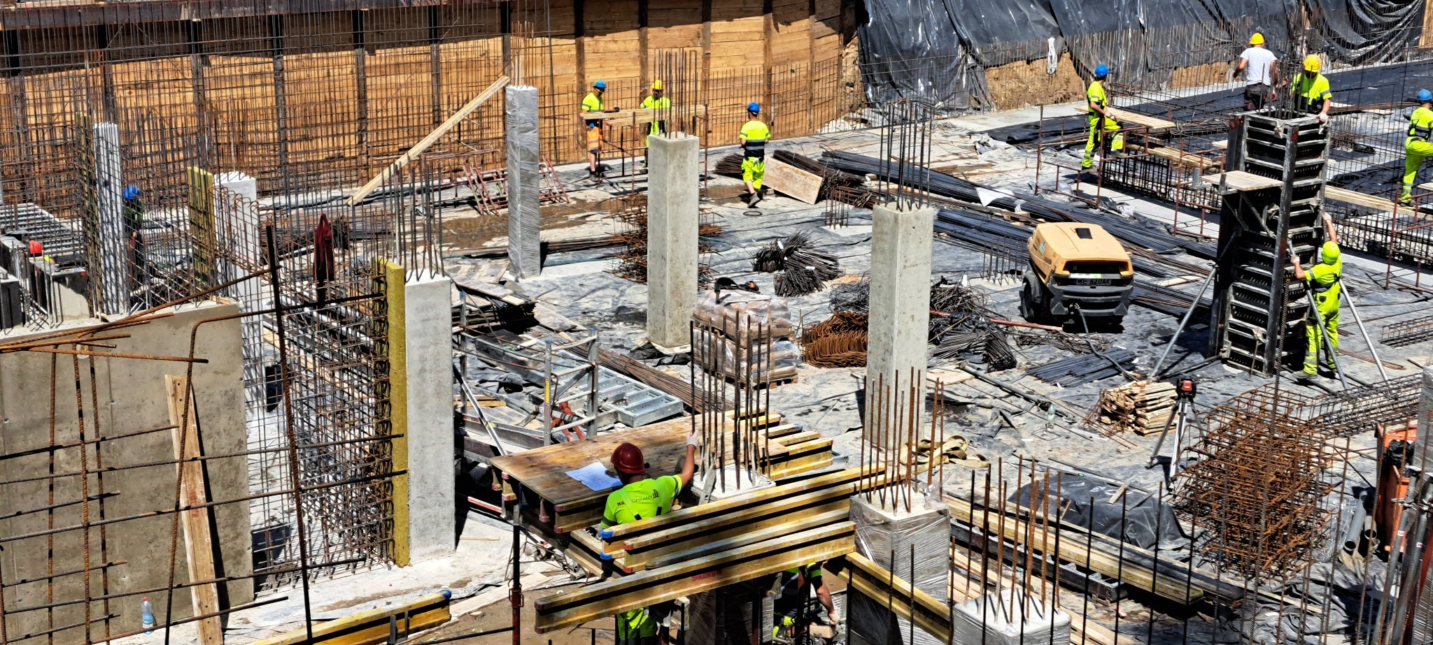 Budowa w toku: robotnicy w żółtych strojach montują szalunki i zbrojenia na placu budowy. Widoczne betonowe słupy, stalowe pręty i maszyny budowlane.