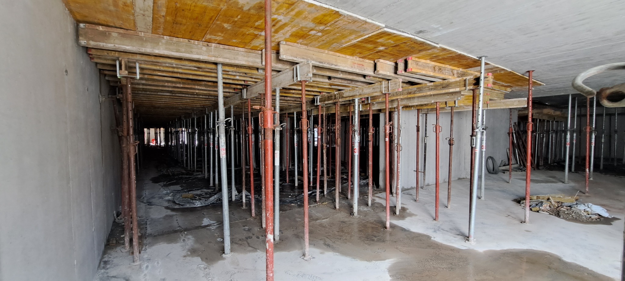 Szare ściany i podpory stropowe w surowym wnętrzu. Widoczne drewniane szalunki stropowe i metalowe stemple podtrzymujące konstrukcję. W tle widoczny otwór.