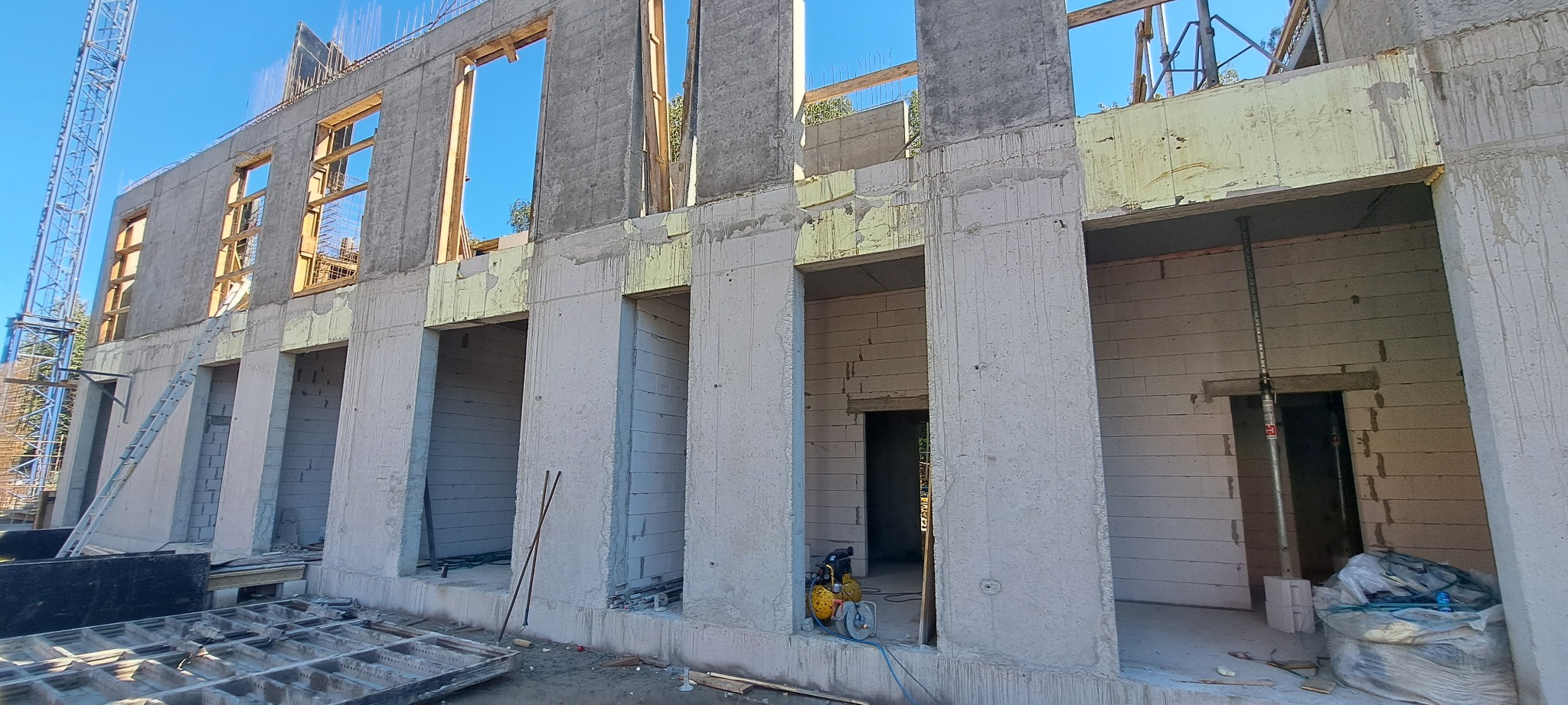 Surowy stan budynku w trakcie budowy z widocznymi otworami okiennymi i drzwiowymi, konstrukcją żelbetową i częściowo wymurowanymi ścianami z bloczków.