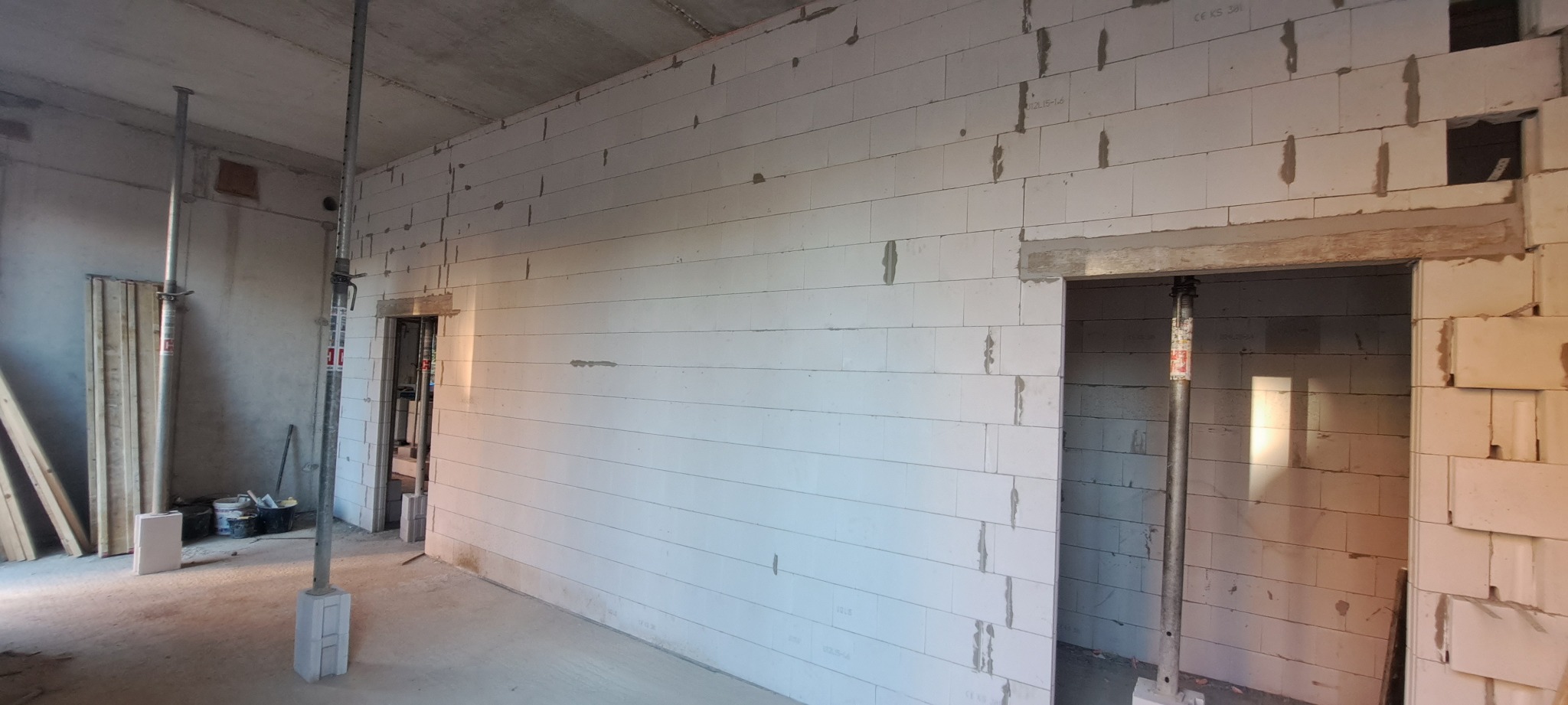 Wnętrze w trakcie budowy: ściany z bloczków, stemple budowlane podtrzymujące nadproża, widoczne narzędzia i materiały budowlane. Stan surowy zamknięty, przygotowanie do dalszych prac.