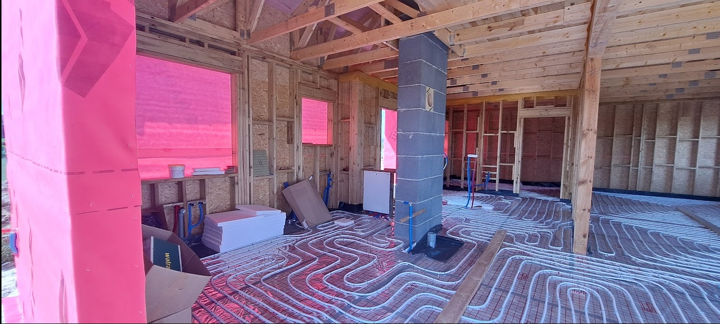 Wnętrze domu w trakcie budowy z ułożonym ogrzewaniem podłogowym, widoczne ściany w konstrukcji szkieletowej i komin z bloczków betonowych. Okna zabezpieczone folią.