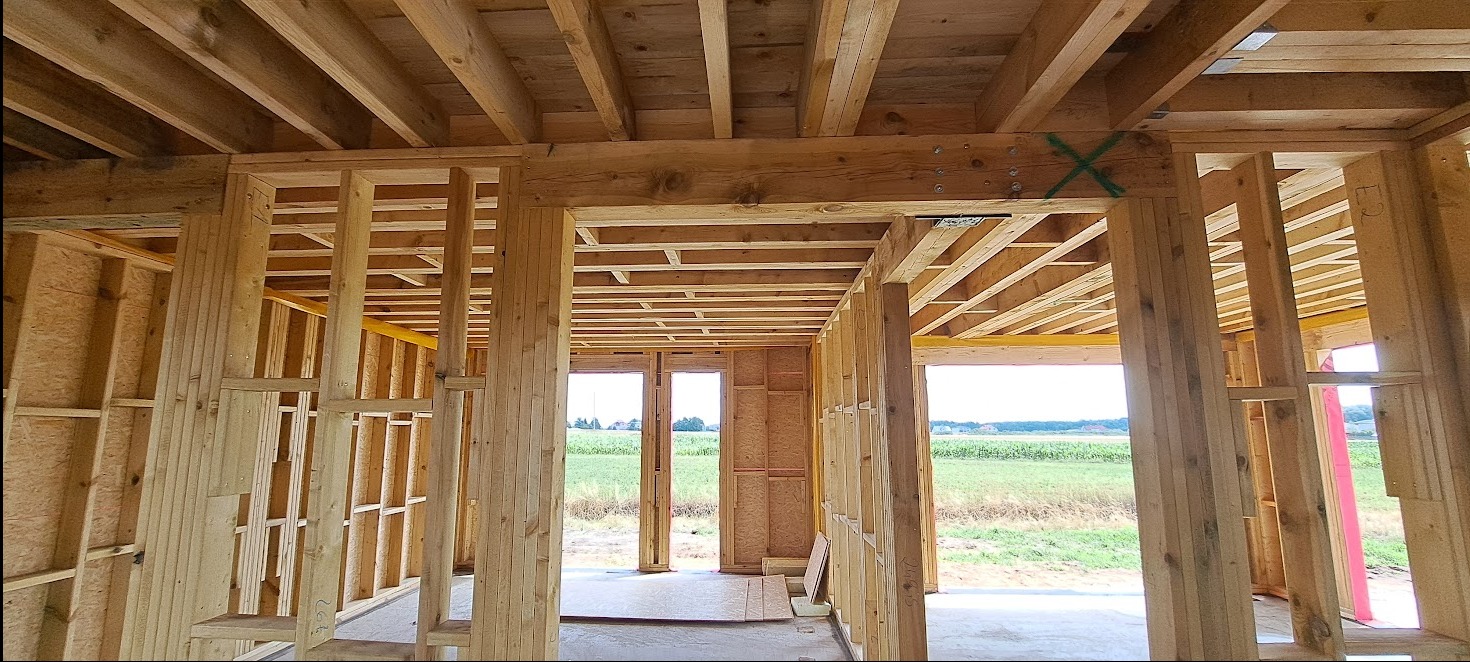 Wnętrze domu w trakcie budowy: drewniany szkielet konstrukcji, widoczne belki stropowe i ściany działowe, zarys okien z widokiem na zielone pole.