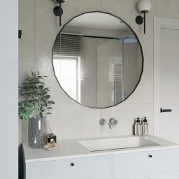 Michał Wilkosiński Conifer Media - Stylowa łazienka z okrągłym lustrem o nieregularnym kształcie, minimalistyczną armaturą i rośliną w szklanym wazonie na blacie umywalki. Widoczna kabina prysznicowa.