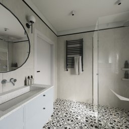 Michał Wilkosiński Conifer Media - Nowoczesna łazienka z okrągłym lustrem, wzorzystą podłogą i szklanym prysznicem. Jasne płytki imitujące marmur, minimalistyczna armatura i designerski grzejnik.