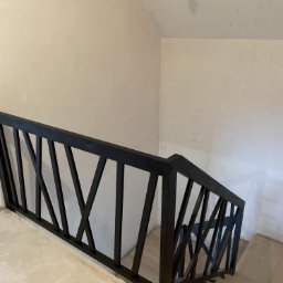 Kokorniak Schody - Nowoczesna, czarna balustrada schodowa z geometrycznym wzorem krzyżujących się belek, zamontowana przy jasnych schodach i ścianach. Minimalistyczny design.