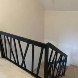 Nowoczesna, czarna balustrada schodowa z geometrycznym wzorem krzyżujących się belek, zamontowana przy jasnych schodach i ścianach. Minimalistyczny design.