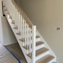 Jasne schody z białymi balustradami i drewnianymi stopniami, widoczne z dołu. Ujęcie podkreśla nowoczesny design i precyzję wykonania.