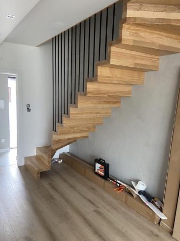 Nowoczesne, wiszące schody drewniane z czarną balustradą w jasnym wnętrzu. Widoczne narzędzia stolarskie. Realizacja we Wschowie.