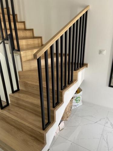 Jasne schody z drewnianymi stopniami i nowoczesną, czarną balustradą. Widok z dołu, podkreślający minimalistyczny design i naturalne wykończenie drewna.