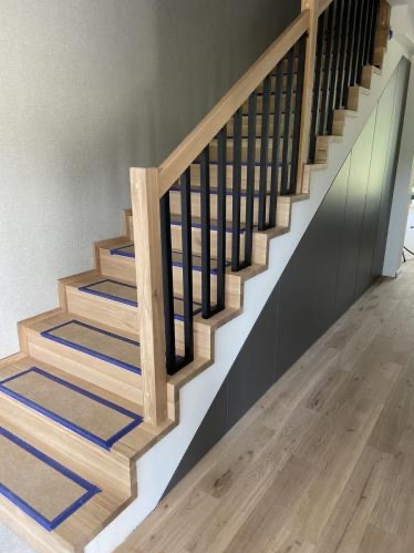 Jasne schody z drewnianymi stopniami zabezpieczonymi folią i czarną, metalową balustradą. Minimalistyczny design, jasne drewno i ciemne akcenty.