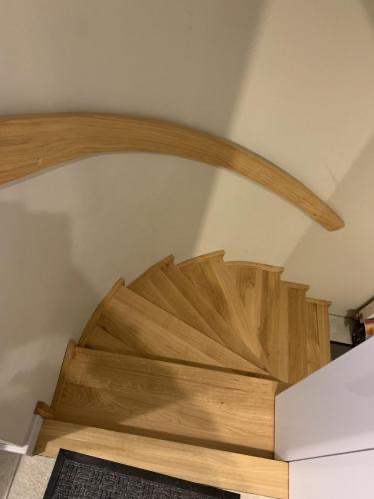 Widok z góry na spiralne schody z jasnego drewna, z zakrzywionym poręczą. Stopnie ułożone w łagodny łuk, szara wycieraczka u dołu. Minimalistyczny design.