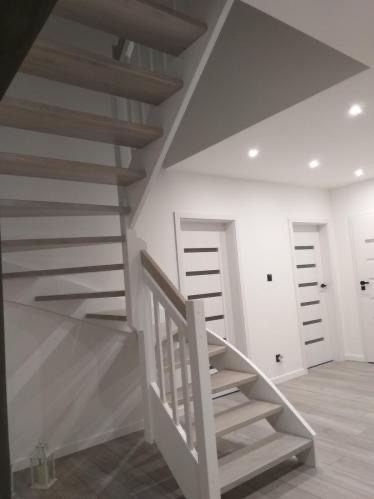 Jasne schody z drewnianymi stopniami i białą balustradą, widok od dołu. W tle białe ściany i trzy białe drzwi z czarnymi paskami. Nowoczesne wnętrze.