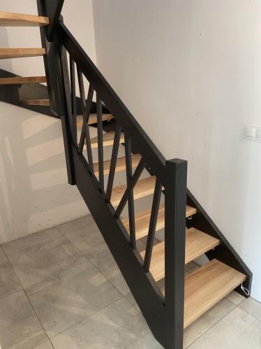 Nowoczesne schody z drewnianymi stopniami i czarną, geometryczną balustradą, widziane z dołu, z delikatnym oświetleniem i szarymi płytkami na podłodze.
