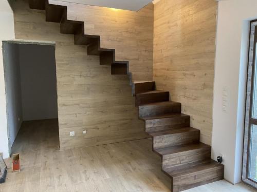 Drewniane schody wspornikowe, zintegrowane ze ścianą wykończoną panelami drewnopodobnymi, w jasnym wnętrzu z widocznym otworem drzwiowym.