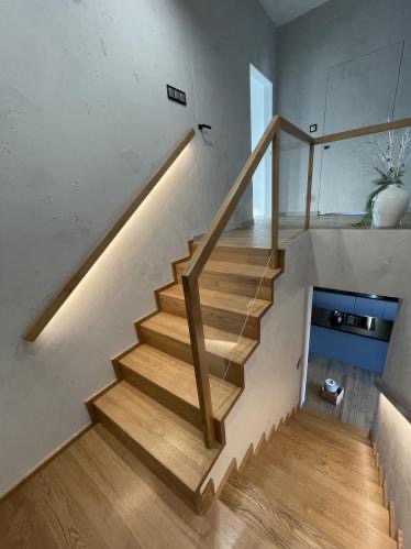 Jasne, drewniane schody z podświetleniem LED i szklaną balustradą w nowoczesnym wnętrzu. Widok z dołu, naturalne światło.