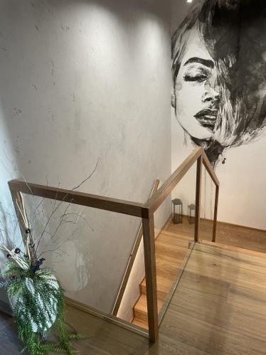 Nowoczesne schody z drewnianymi stopniami i szklaną balustradą, doświetlone subtelnym światłem. Na ścianie widoczny artystyczny mural. Całość tworzy elegancką przestrzeń.