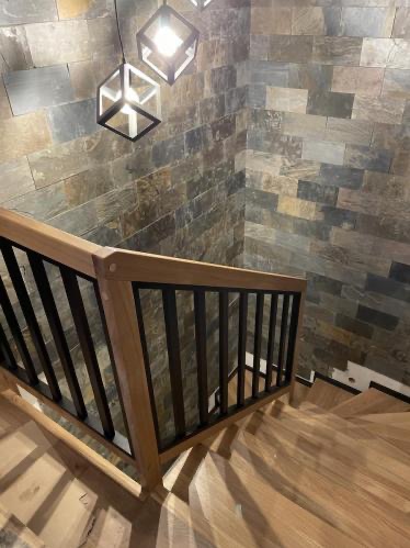 Drewniane schody z czarną balustradą i kamienną ścianą, oświetlone nowoczesnymi lampami. Widok z góry, ukazujący detale wykończenia i unikalny design.