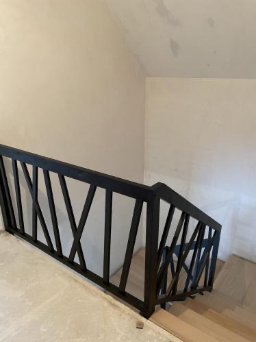Nowoczesna, czarna balustrada schodowa z geometrycznym wzorem krzyżujących się belek, zamontowana przy jasnych schodach i ścianach. Minimalistyczny design.