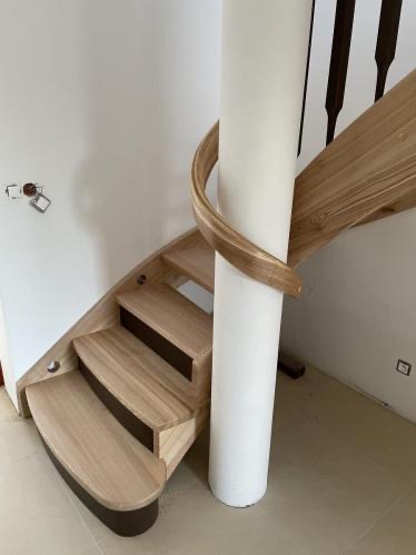 Spiralne schody drewniane z jasnego drewna owijają się wokół białej kolumny. Widoczne gniazdko elektryczne na ścianie i fragment balustrady.